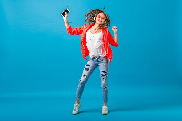 Attractive smiling happy woman dancing listening to music in headphones dressed in hipster style outfit isolated on blue studio background, wearing pink jacket and sunglasses