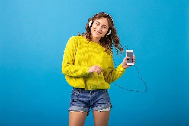 Привлекательная улыбающаяся счастливая женщина танцует, слушая музыку в наушниках в стильном хипстерском наряде, изолированном на синем фоне студии, в шортах и желтом свитере