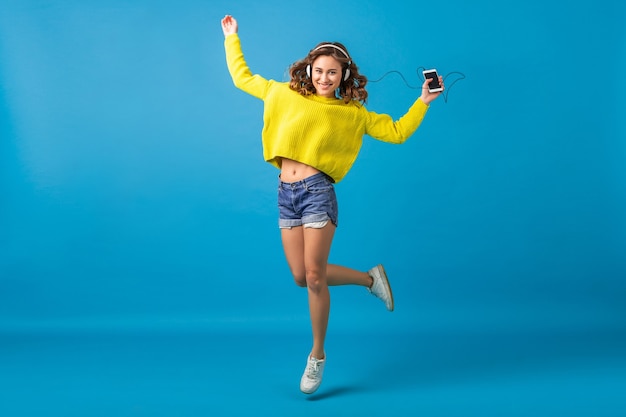 魅力的な笑顔の幸せな女性は、ショートパンツと黄色のセーターを着て、青いスタジオの背景に分離された流行に敏感な衣装でヘッドフォンで音楽を聴いて踊ってジャンプ