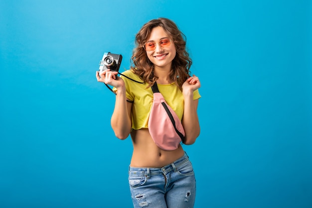 Attractive smiling happy woman posing with vintage photo camera taking pictures dressed in hipster summer colorful outfit isolated on blue studio background