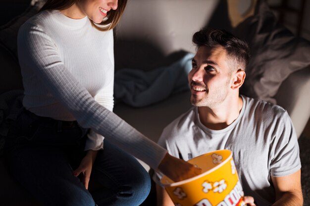 Signora sorridente attraente che prende popcorn dal canestro vicino al positivo ragazzo bello sul sofà