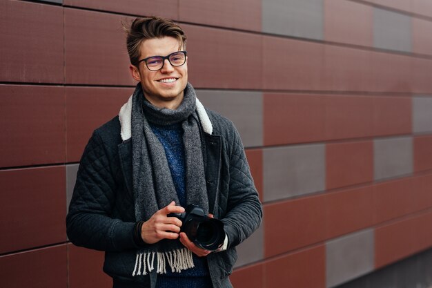 Привлекательный улыбающийся человек в очках проводит профессиональную камеру на улице против стены.