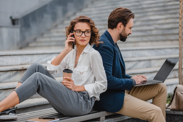 Attractive smiling man and woman talking on phone sitting on stairs in urban city center