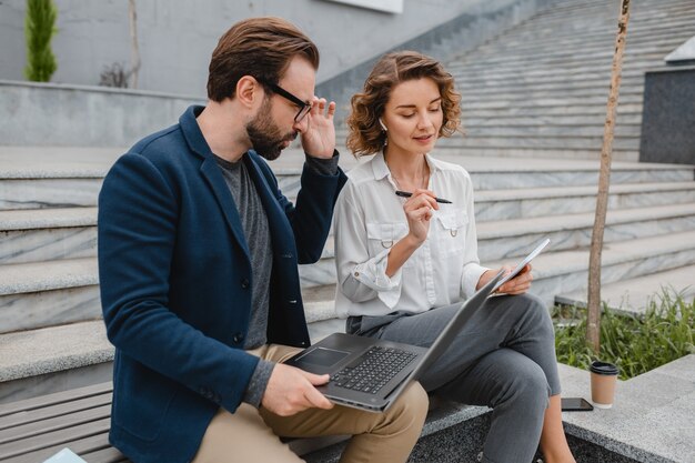 Attractive smiling man and woman talking sitting on bench in urban city center, making notes