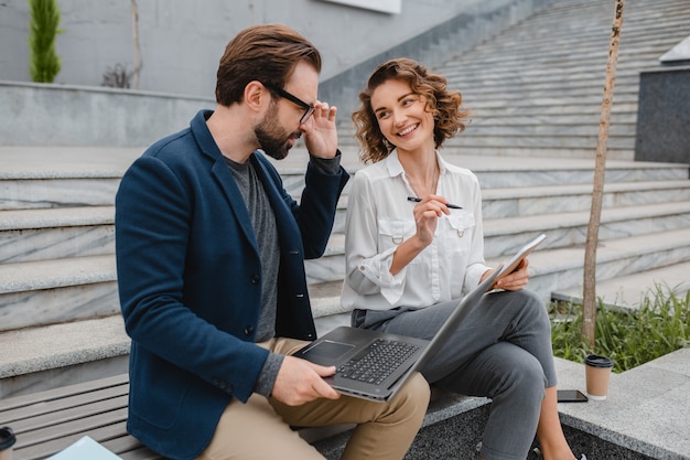 Attractive smiling man and woman talking sitting on bench in urban city center, making notes