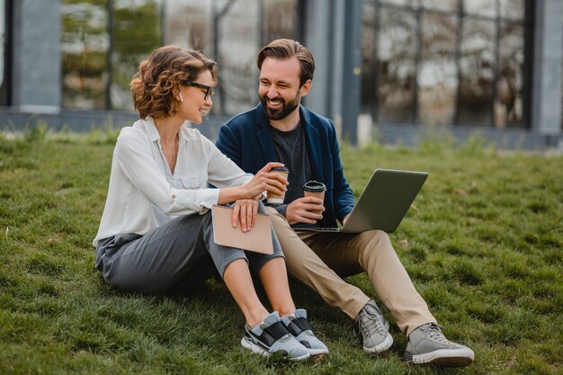 Attractive smiling man and woman talking sitting on grass in urban park, making notes