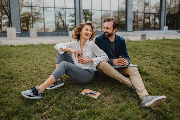 Привлекательный улыбающийся мужчина и женщина разговаривают, сидя на траве в городском парке, делая заметки