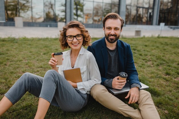 Attractive smiling man and woman talking sitting on grass in urban park, making notes