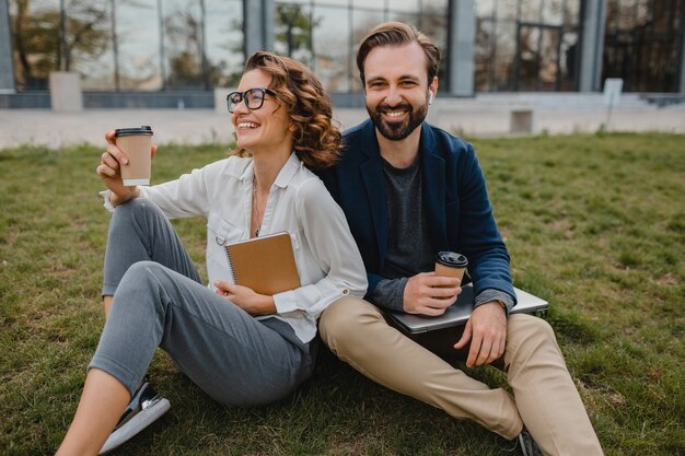 Attractive smiling man and woman talking sitting on grass in urban park, making notes