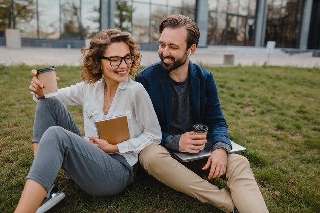 Attractive smiling man and woman talking sitting on grass in urban park, making notes