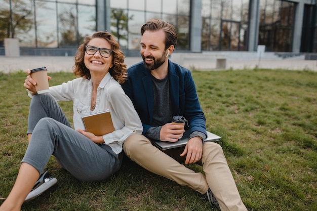 Attractive smiling man and woman talking sitting on grass in urban park, making notes