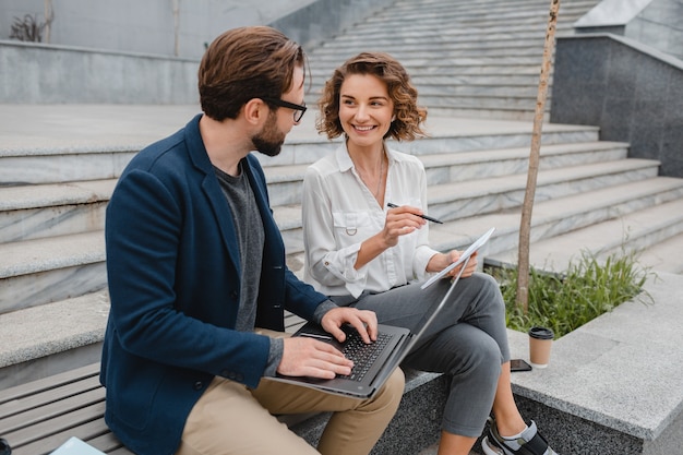 Attractive smiling man and woman talking sitting on stairs in urban city center, making notes