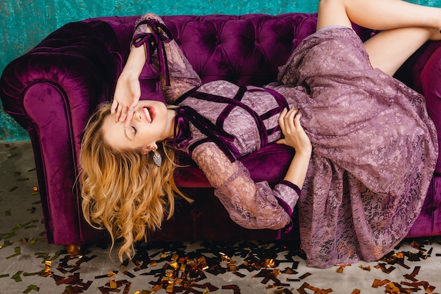 ベルベットのソファの上に横たわるスタイリッシュなバイオレットレースの夜の豪華なドレスで魅力的な笑顔のセクシーな女性