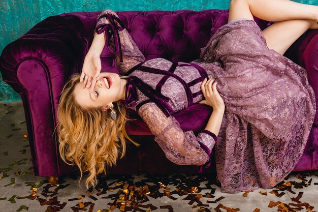 벨벳 소파에 누워 세련된 보라색 레이스 저녁 고급 드레스에 매력적인 웃는 섹시한 여자
