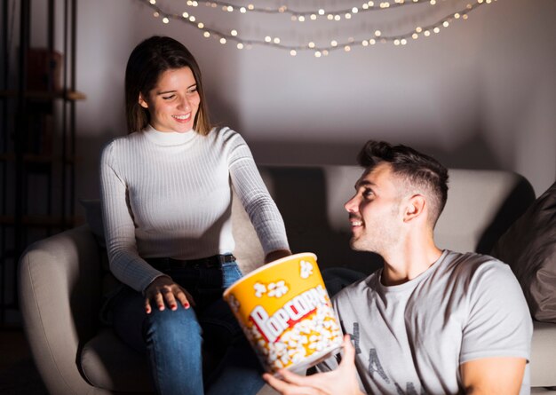 Бесплатное фото Привлекательная женщина улыбается и красивый позитивный человек смотрит телевизор и ест попкорн на диване