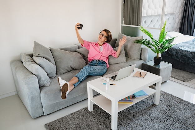 ラップトップの家でオンラインで作業しているテーブルのモダンなインテリアルームの自宅のソファでリラックスして座っているピンクのシャツの魅力的な笑顔の女性