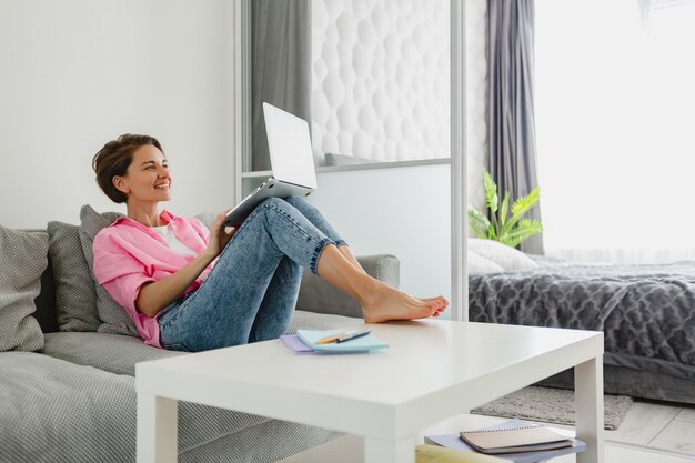 분홍색 셔츠를 입은 매력적인 웃는 여성이 집에서 노트북으로 온라인 작업을 하는 테이블에서 집 소파에 편안하게 앉아 있다