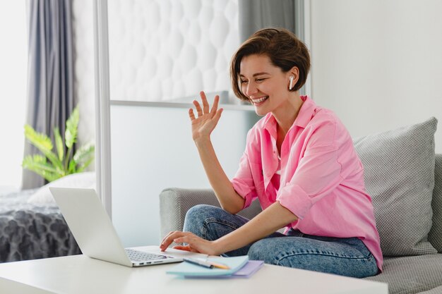 분홍색 셔츠를 입은 매력적인 웃는 여성이 집에서 노트북으로 온라인 작업을 하는 테이블에서 집 소파에 편안하게 앉아 있다