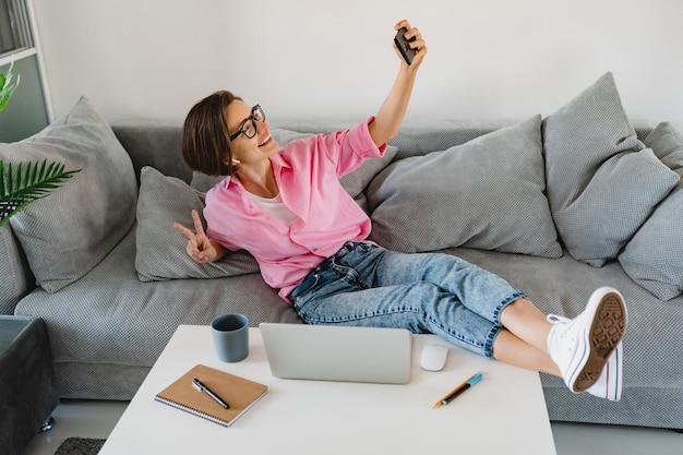 Привлекательная улыбающаяся женщина в розовой рубашке, расслабленно сидящая на диване дома за столом, работающая онлайн