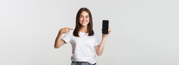 Free photo attractive smiling woman pointing finger at smartphone screen white background