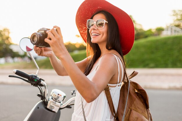 Attractive smiling woman riding on motorbike in street in summer style outfit wearing white dress and red hat traveling on vacation, taking pictures on vintage photo camera