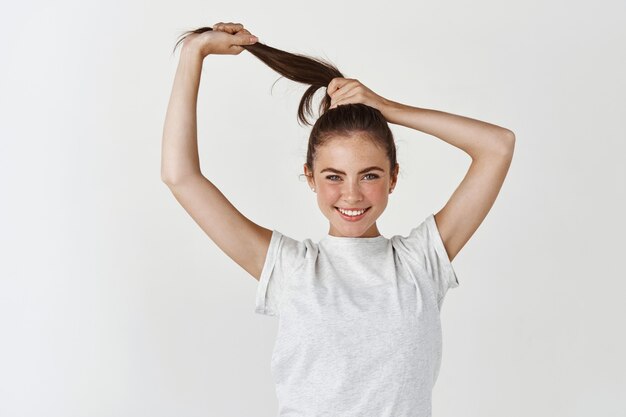 Attractive smiling woman showing long healthy and strong hair, looking sassy at at front, standing over white wall