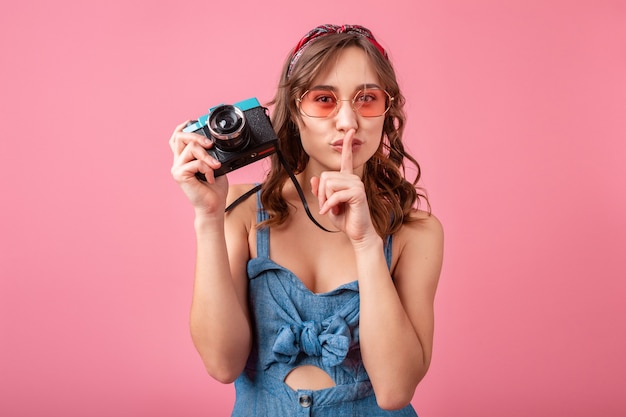 Attractive smiling woman showing silence finger sign taking photo on vintage camera wearing denim dress isolated on pink background