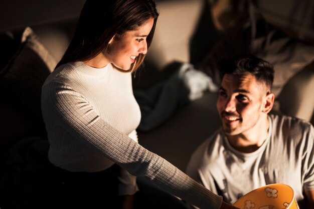 Attractive smiling woman taking popcorn from basket near handsome positive man on sofa