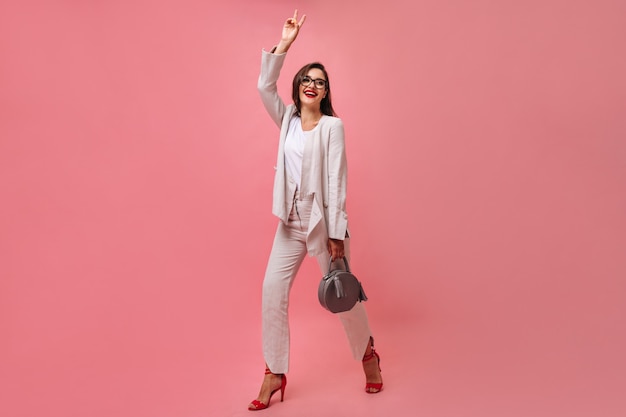 Привлекательная улыбающаяся женщина в белом современном костюме и очках держит сумочку и показывает знак мира на розовом изолированном фоне.