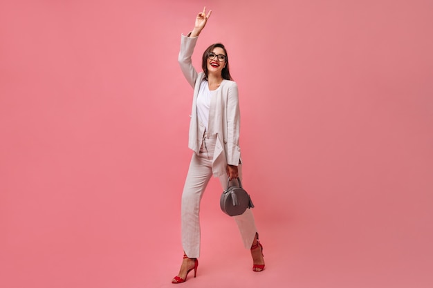 Attractive smiling woman in white modern suit and eyeglasses holds handbag and shows peace sign on pink isolated background.