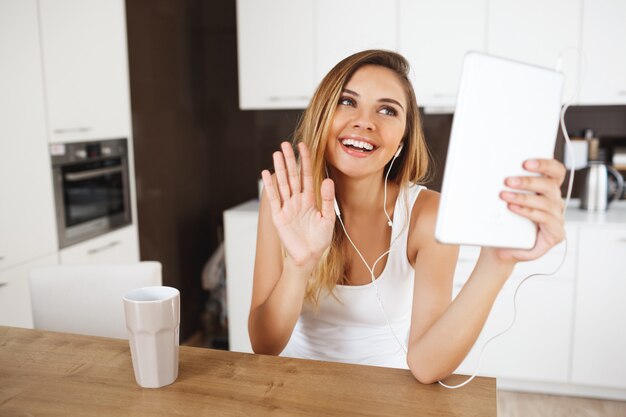 Attractive smiling young girl sitting at dinner table holding tablet and talking with friends via messenger