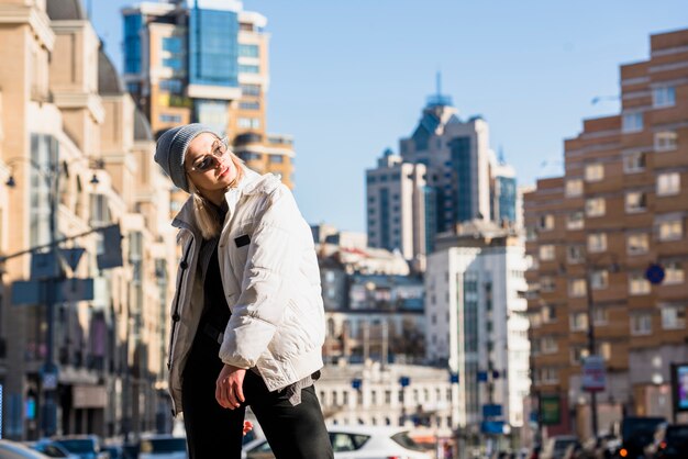 An attractive smiling young woman wearing glasses and jacket posing in front of buildings