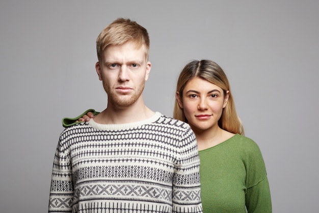 Attractive smiling young woman with blonde hair standing behind serious unshaven man, holding hand on his shoulder as sign of support, standing at grey wall with copy space for your information