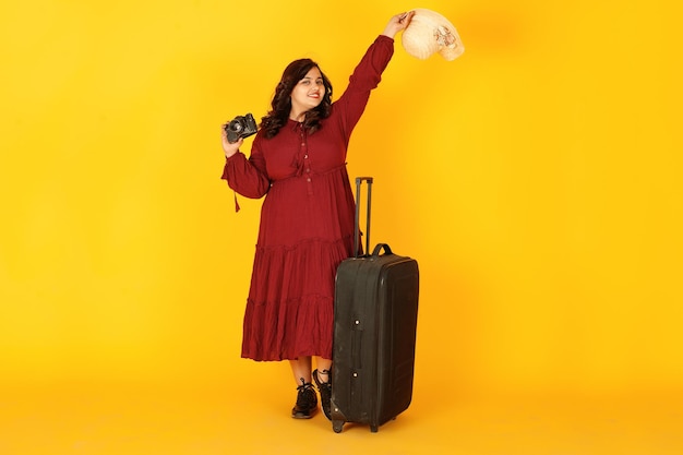 Free photo attractive south asian traveler woman in deep red gown dress hat posed at studio on yellow background with suitcase amd old vintage photo camera