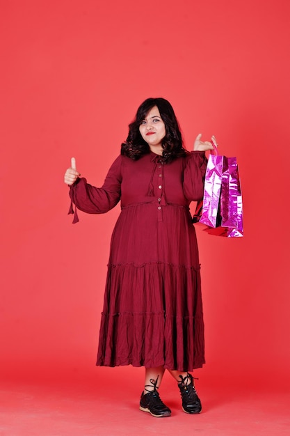 Attractive south asian woman in deep red gown dress posed at studio on pink background with bright colored shopping bags