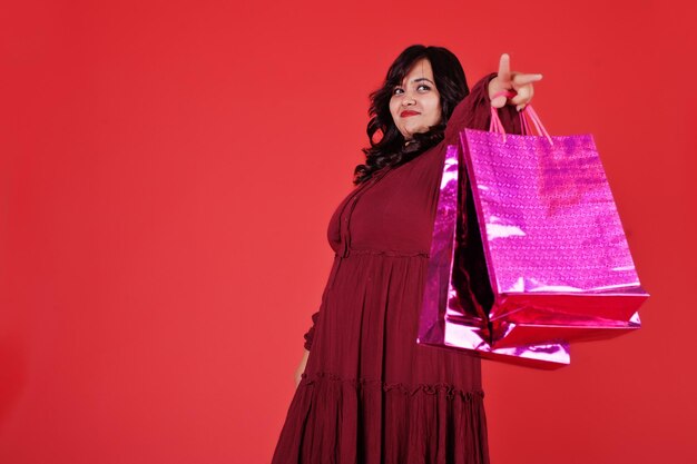 밝은 색의 쇼핑백을 들고 분홍색 배경에 스튜디오에서 포즈를 취한 진한 빨간색 가운 드레스를 입은 매력적인 남아시아 여성
