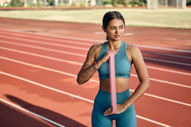 Attractive sporty girl in stylish sportswear stretching hands with rubber band during training on city stadium