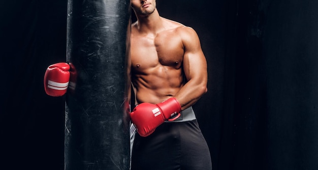 Attractive sporty man is posing with punching bag in dark photo studio.