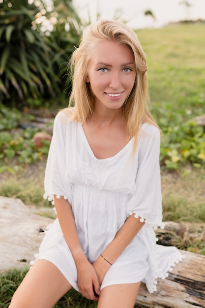 Attractive sporty woman with long blond hair, big blue eyes and clean skin dressed in white dress sitting on wood