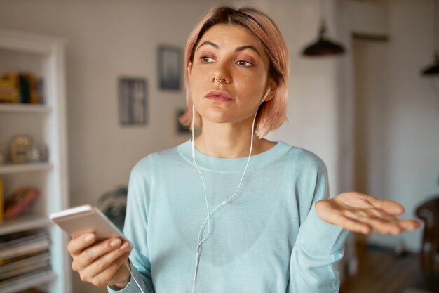 Attractive student girl using earbuds and mic set while communicating online with friend via video chat on smart phone, discussing plans, gesturing.