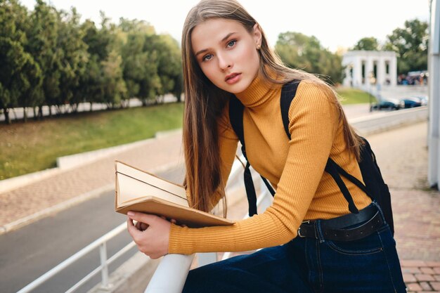 Attractive student girl with text book thoughtfully looking in camera studying in city park