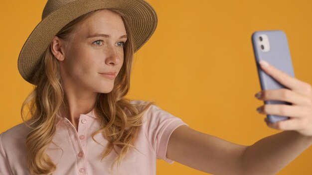 Attractive stylish blond girl in hat taking selfie on smartphone over yellow background