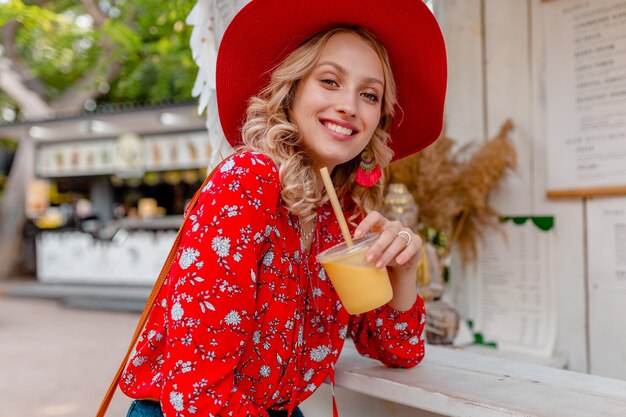 わらの赤い帽子とブラウスの夏のファッション衣装でナチュラルフルーツカクテルスムージーを飲む魅力的なスタイリッシュな金髪の笑顔の女性