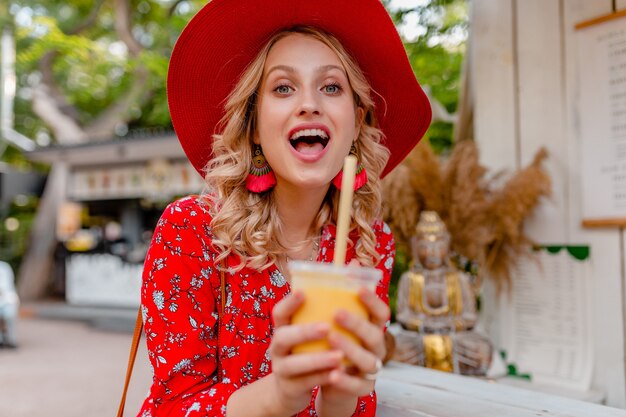 わらの赤い帽子とブラウスの夏のファッション衣装でナチュラルフルーツカクテルスムージーを飲む魅力的なスタイリッシュな金髪の笑顔の女性