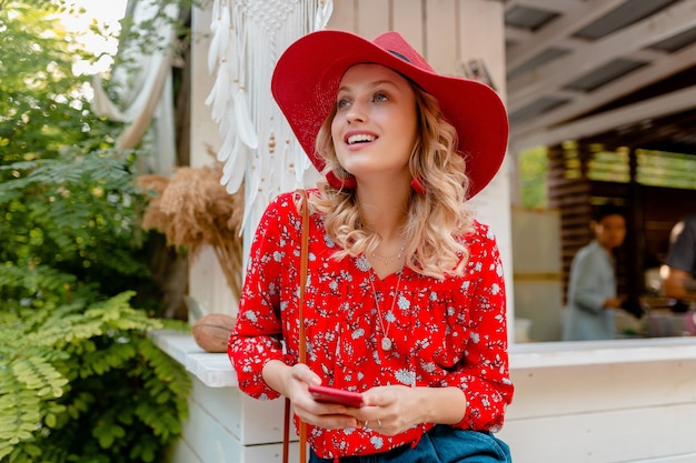 魅力的なスタイリッシュな金髪の笑顔の女性は、スマートフォンのカフェを使用して保持しているわらの赤い帽子とブラウスの夏のファッション衣装で