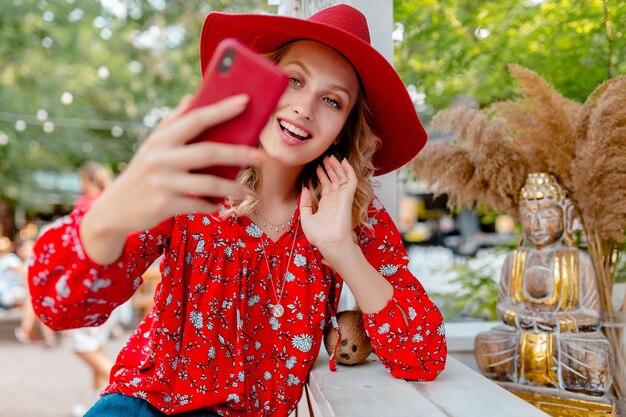 Attractive stylish blond smiling woman in straw red hat and blouse summer fashion outfit taking selfie photo on camera smart phone  cafe