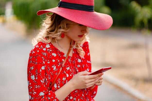 Attractive stylish blond smiling woman in straw red hat and blouse summer fashion outfit  using phone