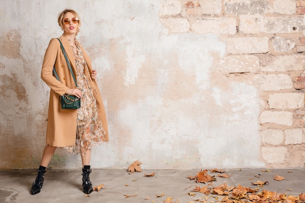 Attractive stylish blonde woman in beige coat posing against vintage wall