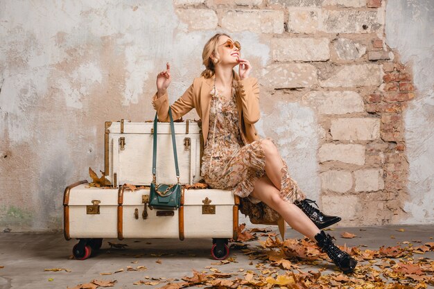 Attractive stylish blonde woman in beige coat sitting on suitcases against wall in street