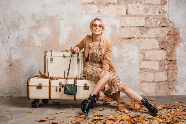 Attractive stylish blonde woman in beige coat sitting on suitcases against wall in street